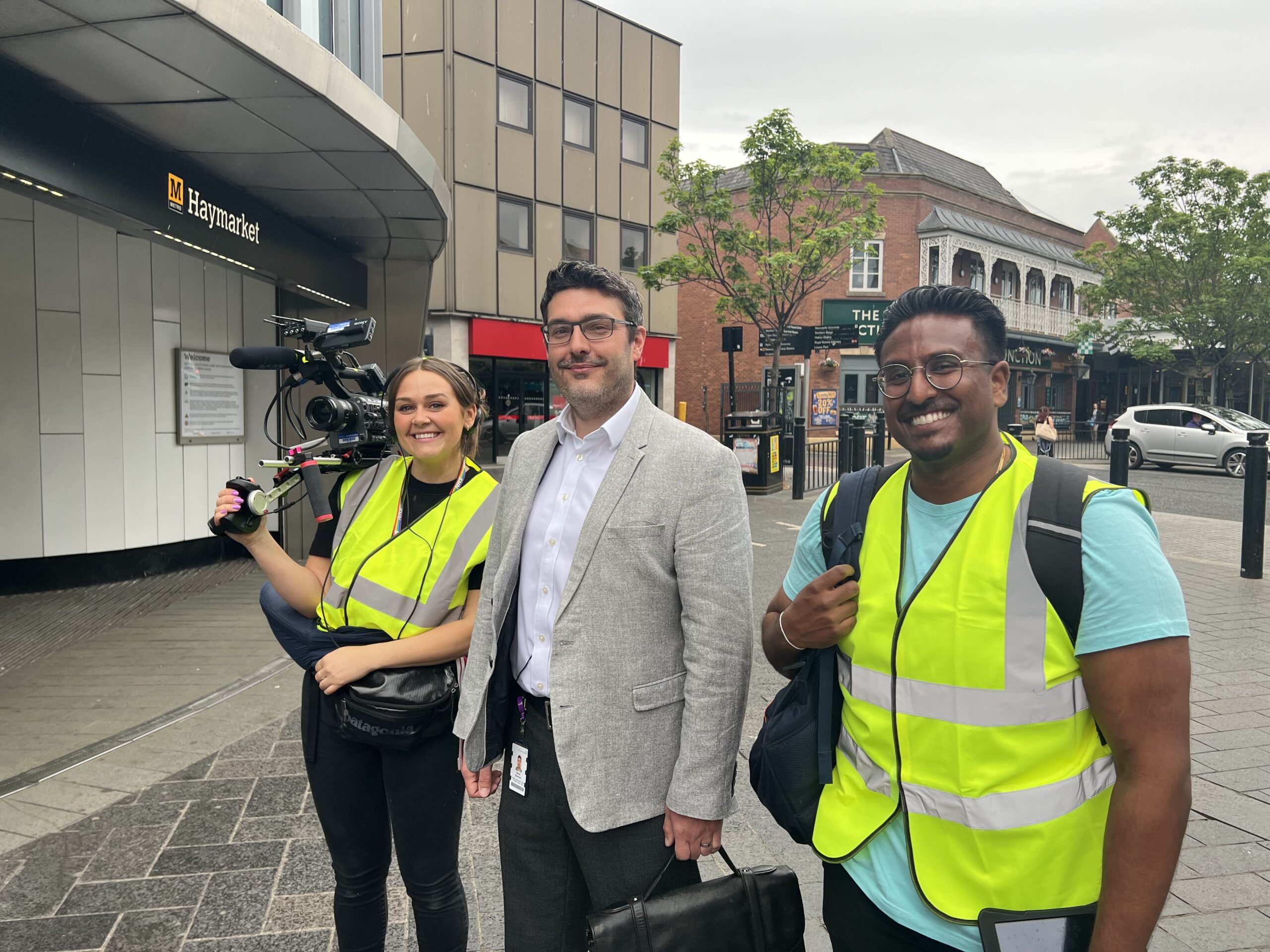 A photo of two people wearing high vis vests and one man wearing a suit outside Haymarket Metro station. One person is carrying a professional camera.